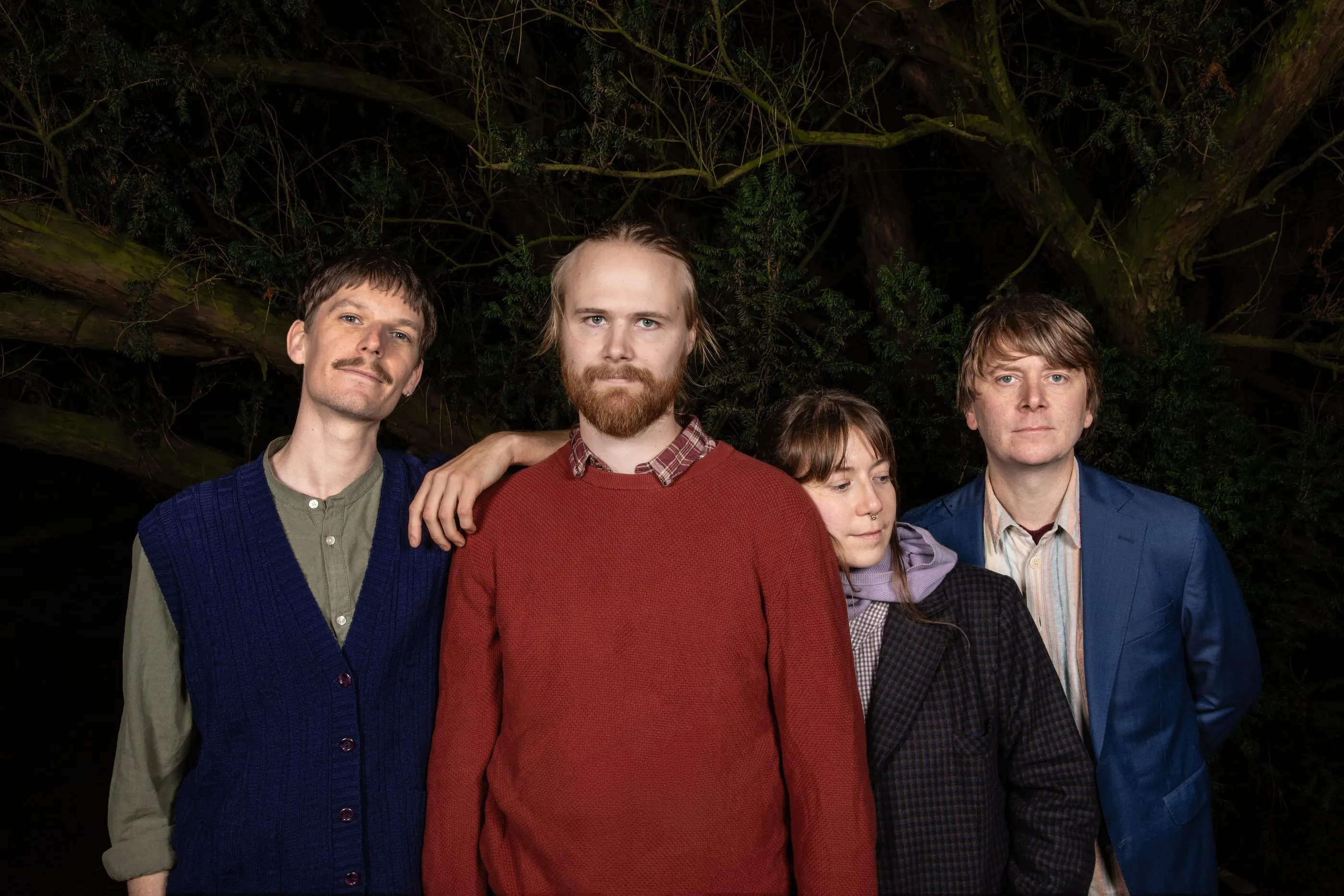 The band standing close together in front of a tree at night.